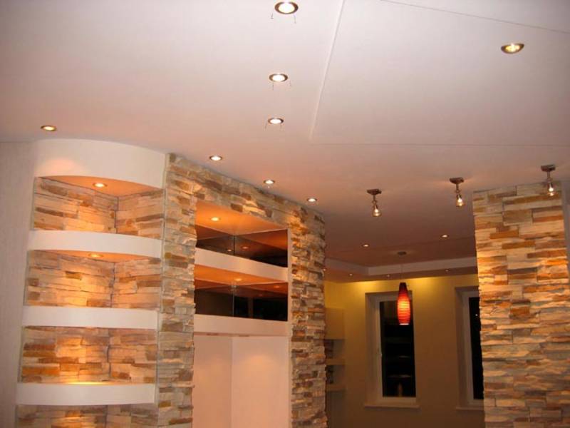 Wykończone wnętrze z dekoracyjną ścianą z kamienia, podświetlanymi półkami i punktowym oświetleniem sufitowym, uzupełnione wiszącą lampą z czerwonym kloszem.